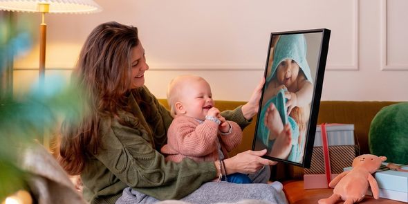 Femme assise dans un salon avec un bébé sur les genoux, tenant une décoration murale personnalisée du bébé que celui-ci regarde en riant, pour illustrer les idées de cadeaux photo de Photobox pour le premier Noël de bébé.