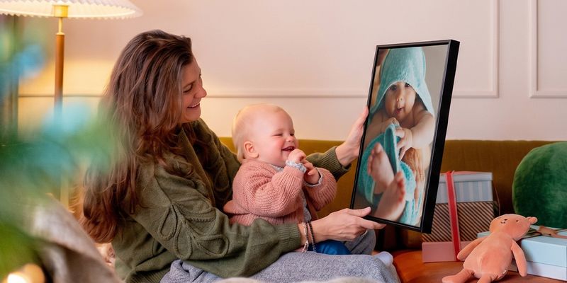 Femme assise dans un salon avec un bébé sur les genoux, tenant une décoration murale personnalisée du bébé que celui-ci regarde en riant, pour illustrer les idées de cadeaux photo de Photobox pour le premier Noël de bébé.