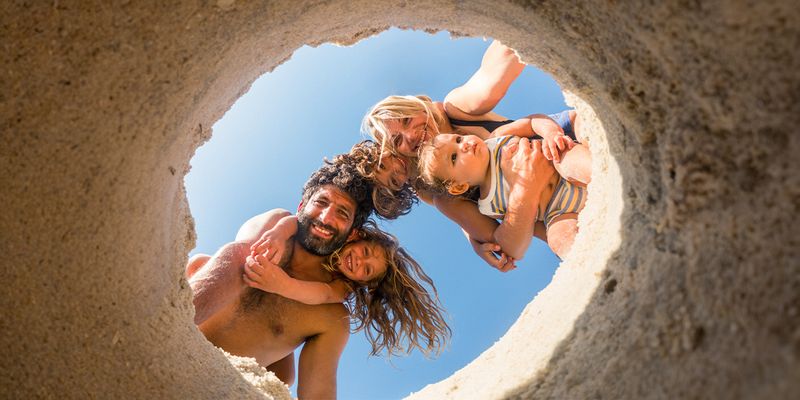 Une famille de quatre personnes à la plage – maman, papa, une petite fille et un petit garçon – regarde un trou creusé dans le sable, capturant un moment de jeu d’été et inspirant des idées de photos de plage.