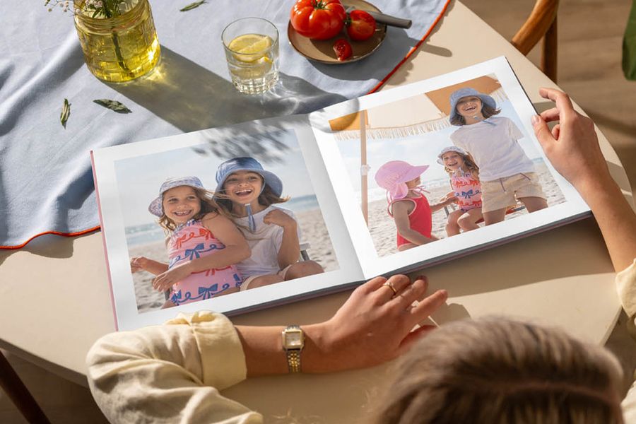 Un livre photo ouvert sur une table de cuisine présente des images de jeunes filles s'amusant à la plage, illustrant le conseil de Photobox pour se replonger dans les souvenirs de l'été grâce à un livre photo personnalisé.