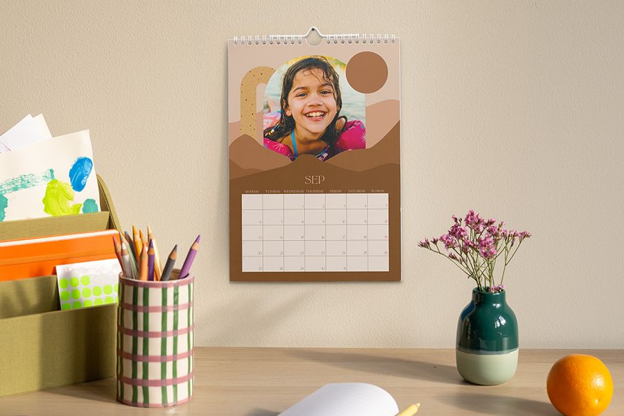 Un calendrier personnalisé affichant le mois de septembre et une photo d'une jeune fille est accroché au-dessus d'un bureau avec un pot à crayons, un vase de fleurs et un organiseur contenant des fournitures créatives et scolaires, illustrant le conseil de Photobox pour créer un espace de travail propice à la concentration pendant l'année scolaire.