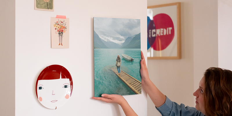 Une femme accroche une décoration murale personnalisée sur son mur, à côté d'une assiette décorative et de quelques tirages photo, présentant le guide étape par étape de Photobox pour accrocher sa décoration murale avec les kits d'accrochage.