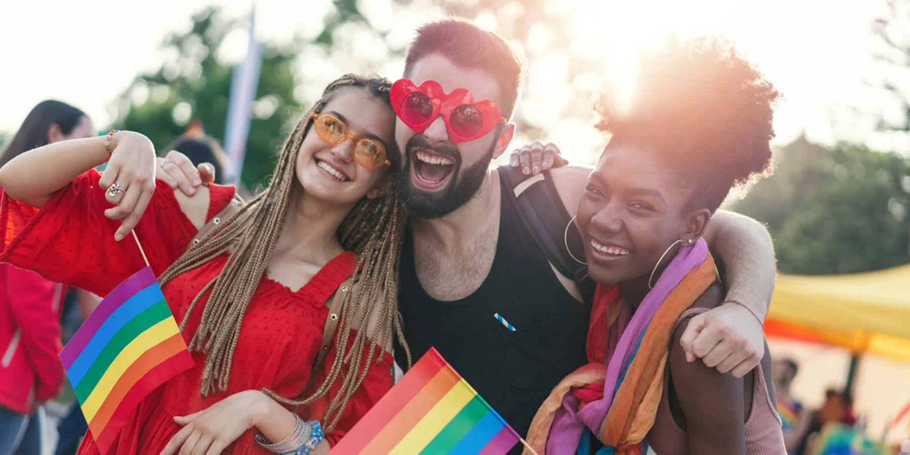 Trois amis sourient lors d’un événement Pride, tenant des drapeaux arc-en-ciel et portant des lunettes de soleil, célébrant la joie et l’unité de la Pride LGBTQI+.
