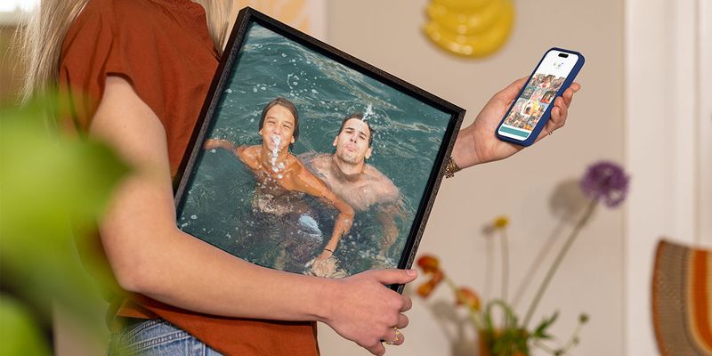 Une femme tient sous son bras une photo de vacances encadrée montrant un homme et un jeune garçon jouant dans l'eau, tout en sélectionnant d'autres photos sur son téléphone, illustrant le guide de Photobox pour transformer ses photos de vacances en décoration murale.