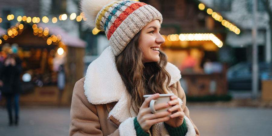 Femme vêtue d’un manteau douillet et d’un bonnet, tenant une tasse dans un marché de Noël, avec des lumières festives et des chalets en arrière-plan, pour illustrer le guide de Photobox sur les plus beaux marchés de Noël d’Europe.