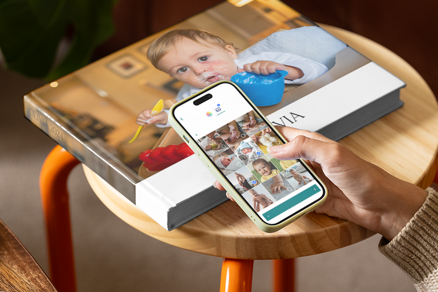Une main faisant défiler des photos de bébé sur un smartphone avec un mon premier album photo de Photobox sur une table en bois à l'arrière-plan.