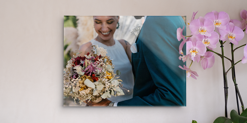 Une photo mariage cadre de Photobox montrant une mariée souriante avec son bouquet, posée sur un mur à côté d'une orchidée.