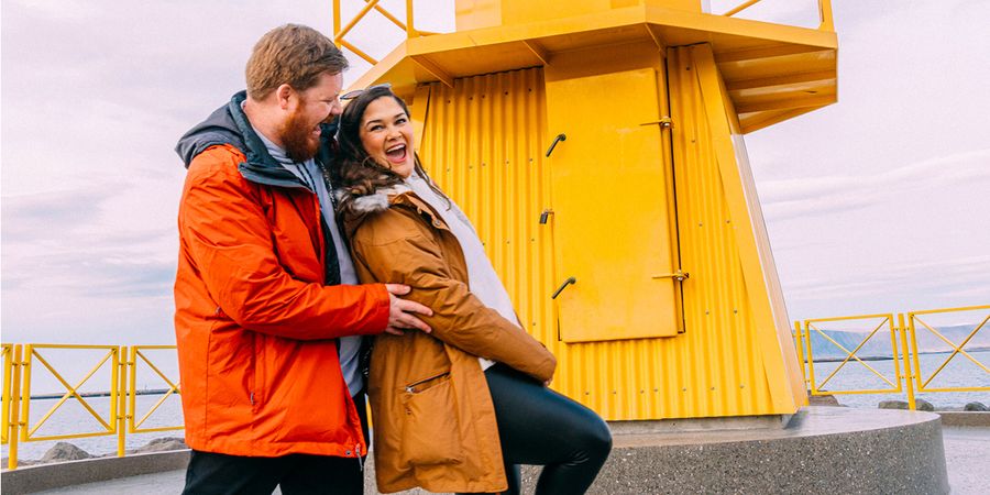Un couple heureux qui rit devant un phare jaune en cherchant quoi écrire pour la Saint Valentin et une idée cadeau Saint Valentin pour accompagner leurs créations Photobox.