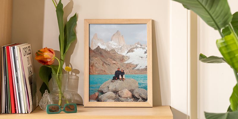 Une photo de paysage encadrée posée sur une étagère en bois à côté de livres et de fleurs, illustrant comment décorer sans percer avec Photobox.