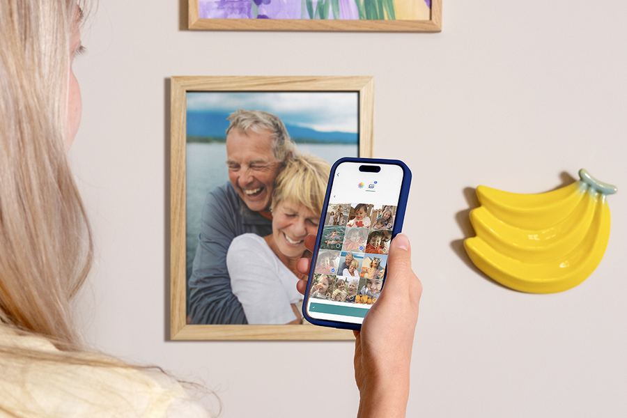 Une femme tient son téléphone pour montrer comme il est facile de commander des tirages photo Photobox depuis l’application, avec des tirages photo encadrés sur le mur derrière elle, pour inspirer le tirage photo en ligne et l’impression de photos depuis son téléphone.