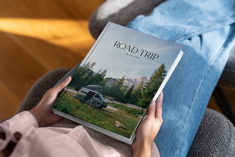 Un beau livre photo Photobox intitulé Road Trip repose sur les genoux de quelqu’un, avec un paysage de forêt et un camping-car en couverture, pour inspirer un livre photo de voyage décoratif.