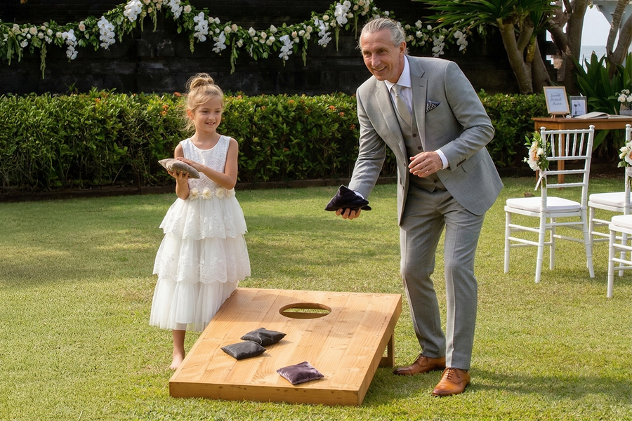 Une jeune fille en tenue élégante de mariage et un homme plus âgé en costume jouent au cornhole lors d’un mariage en lançant des sacs dans la cible, pour inspirer Photobox et des jeux mariage en plein air.