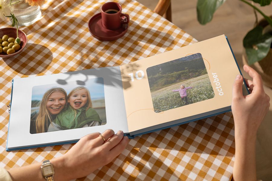 Un livre photo Photobox de type beau livre ouvert, montrant une mère et sa fille sur une page et une petite fille qui gambade dans un champ de fleurs sur l’autre, pour inspirer un livre photo décoratif personnalisé et un album photo de famille pour table basse.