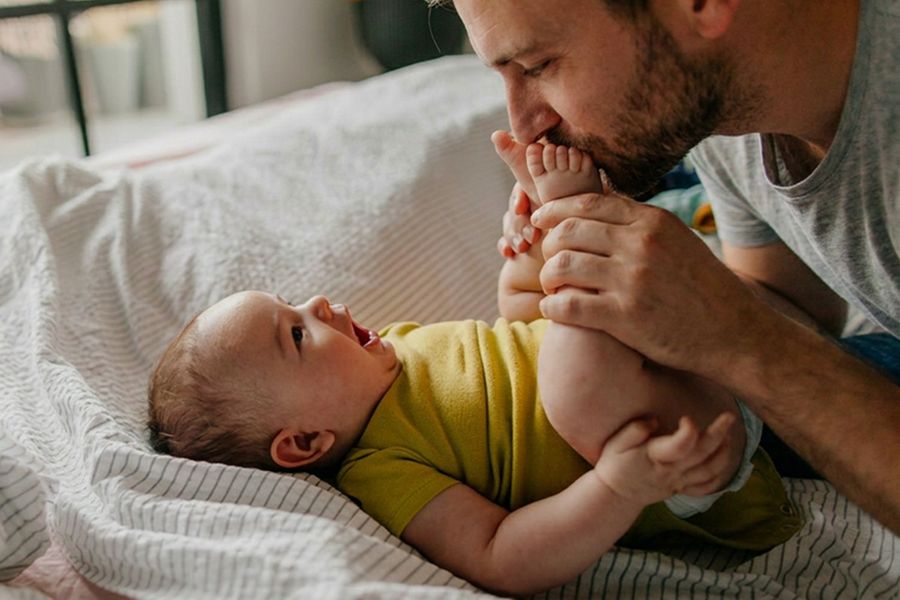 Un père embrasse les pieds de son bébé, pour inspirer une carte fête des pères pour une première fête des pères et un message fête des pères plein de tendresse.