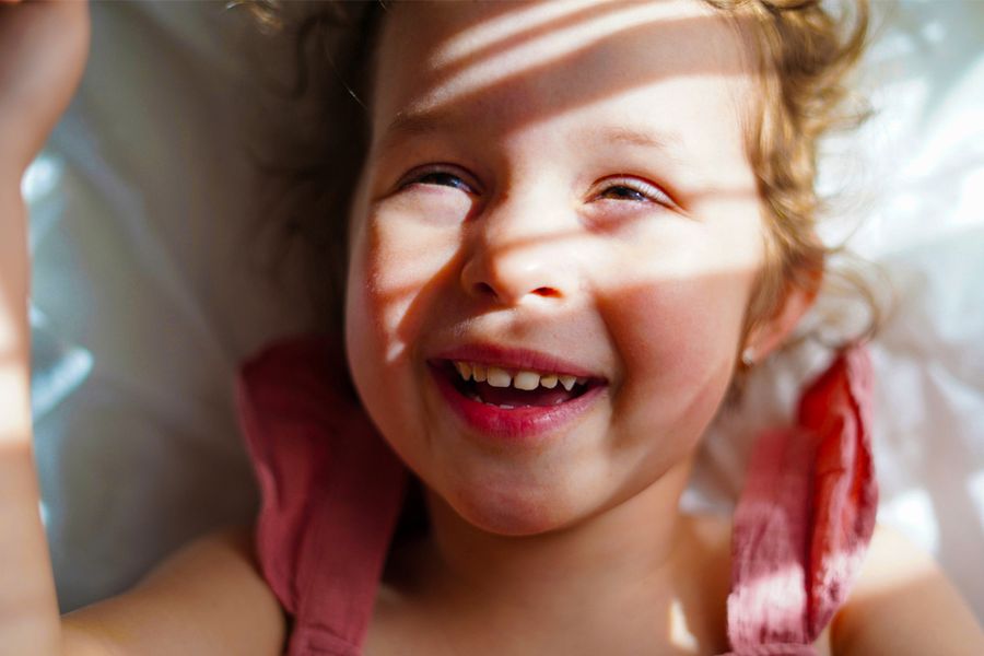Une petite fille souriante avec le soleil et des ombres douces sur le visage, pour inspirer Photobox et apprendre à prendre de belles photos au soleil.