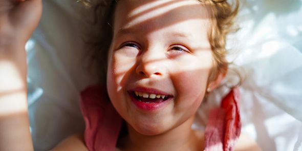 Une petite fille souriante avec le soleil et des ombres douces sur le visage, pour inspirer Photobox et apprendre à prendre de belles photos au soleil.