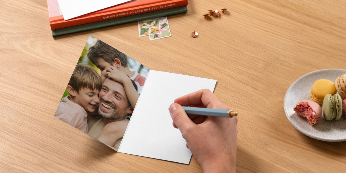 Une carte photo personnalisée Photobox pour la fête des pères est ouverte sur une table et montre un père avec ses deux fils, avec un stylo prêt à écrire un message à côté de livres et d’une assiette de macarons, pour inspirer un message de fête des pères.
