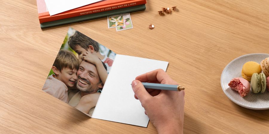 Une carte photo personnalisée Photobox pour la fête des pères est ouverte sur une table et montre un père avec ses deux fils, avec un stylo prêt à écrire un message à côté de livres et d’une assiette de macarons, pour inspirer un message de fête des pères.