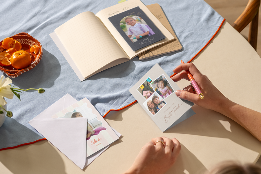 La main d’une femme repose sur une table, prête à écrire dans une carte de remerciement maîtresse personnalisée Photobox illustrée de plusieurs photos d’enfants souriants.