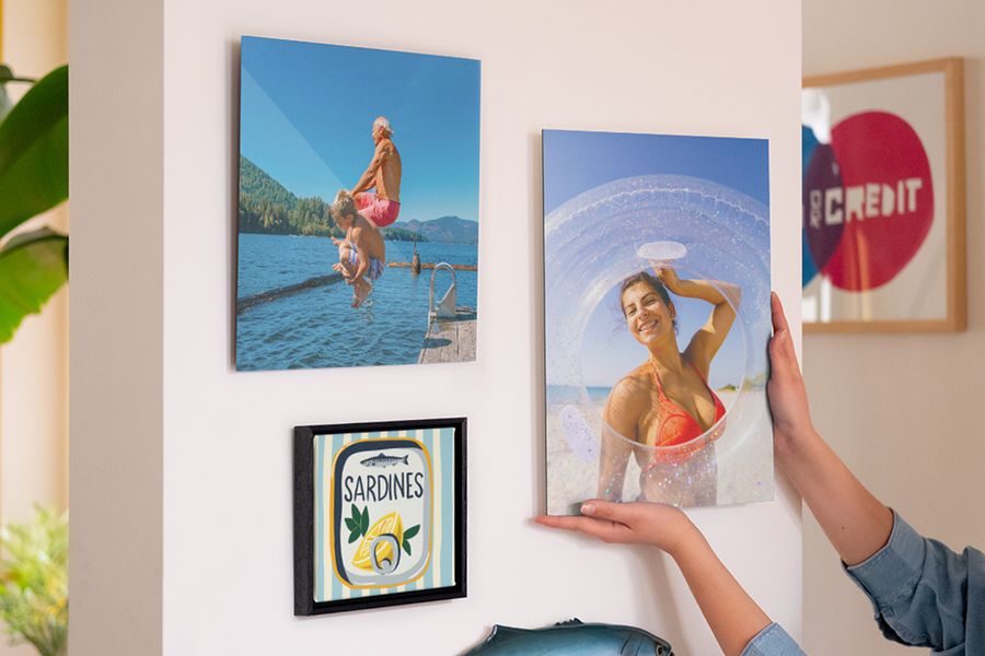 Un mur de cadres avec des décorations murales personnalisées Photobox montre une femme souriante à la plage, un homme et un garçon sautant dans la mer, ainsi qu’une illustration de boîte de sardines, pour inspirer une déco été lumineuse et pleine de fraîcheur.