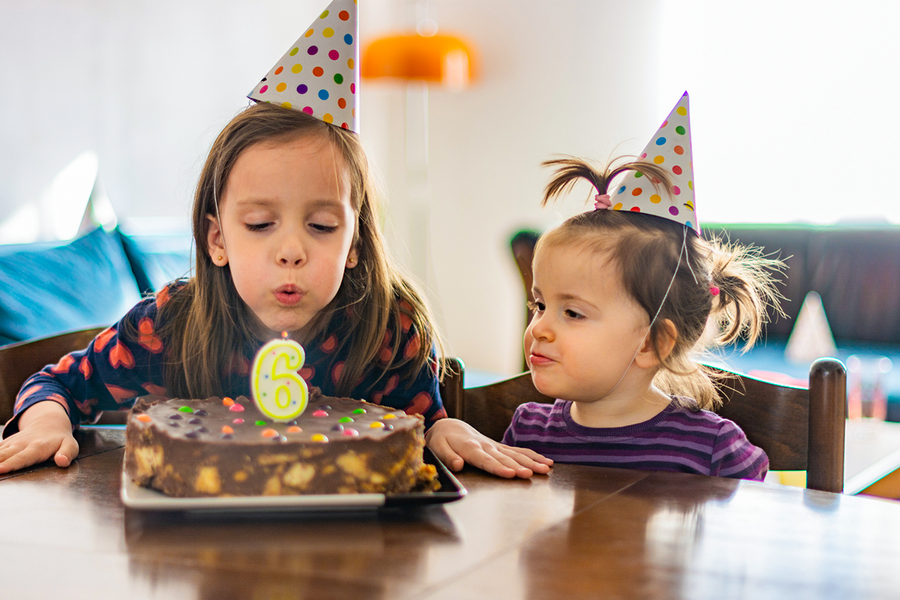 Une petite fille avec un chapeau de fête soufflant sa bougie de 6ème anniversaire avec une amie, illustrant une idée cadeau anniversaire enfant de Photobox.