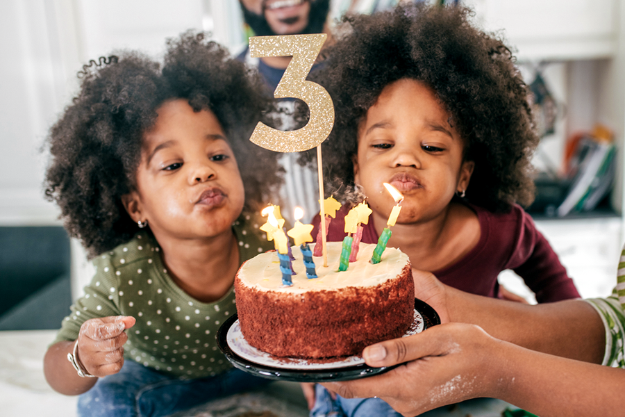 Deux petites filles jumelles s'apprêtant à souffler les bougies de leur gâteau de 3ème anniversaire, illustrant une idée cadeau anniversaire ou un cadeau personnalisé anniversaire de Photobox.