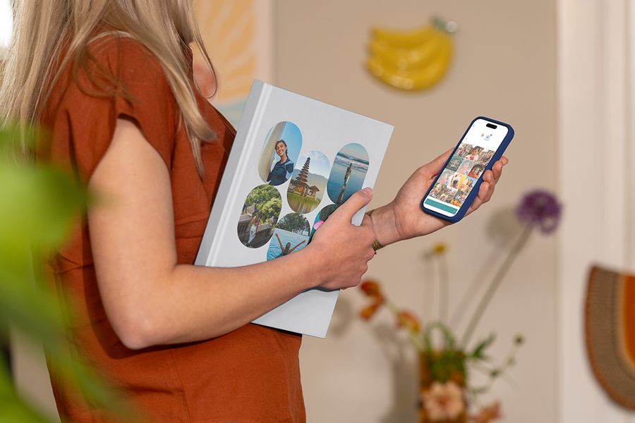 Une femme tient un album photo vacances Photobox fermé avec des photos de voyage en famille sur la couverture tout en regardant des images de voyage sur son téléphone, illustrant comment choisir les bonnes photos pour un livre photo voyage personnalisé.