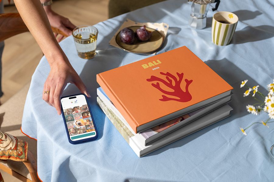 Une pile de différents livres photo voyage Photobox est posée à côté d’une main tenant un téléphone avec une photo de vacances prête à être ajoutée, avec en haut un livre photo Bali 2026 montrant du corail sur la couverture, illustrant comment créer un album photo voyage personnalisé.