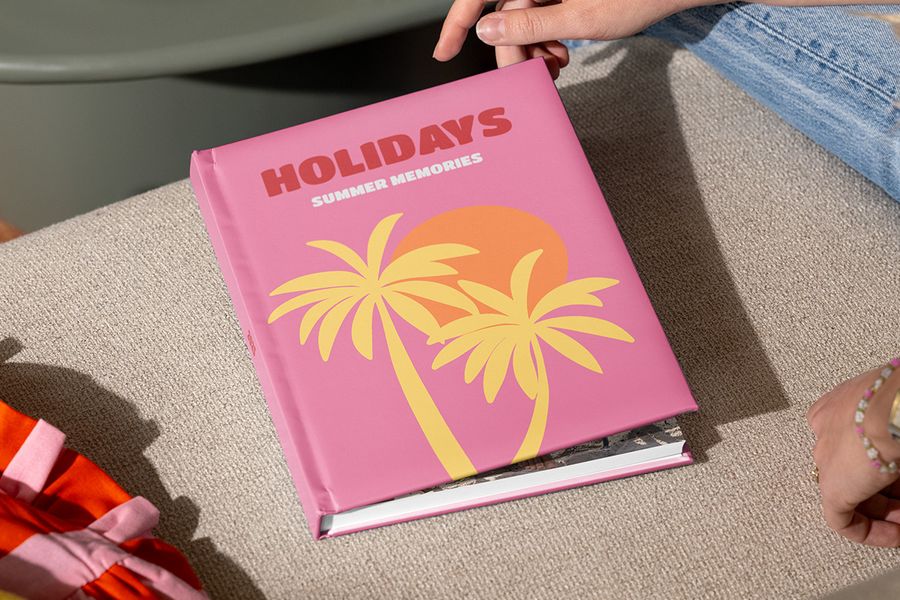 Un livre photo voyage Photobox fermé intitulé « Holidays, summer memories », avec des palmiers illustrés sur une couverture rose, est tenu par des mains prêtes à l’ouvrir, illustrant comment structurer un album photo vacances et raconter ses souvenirs de voyage.