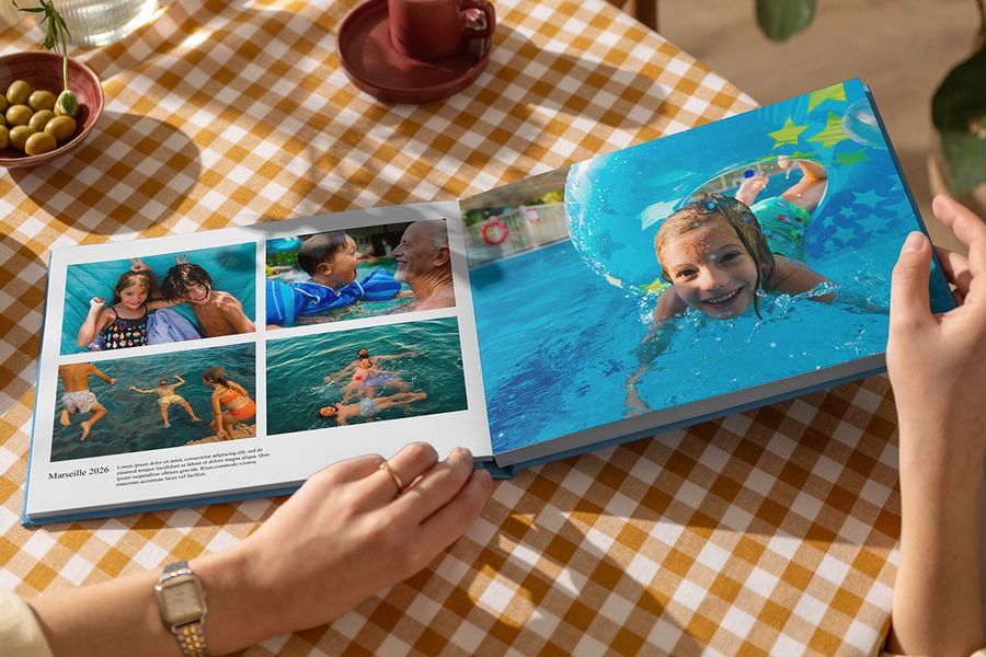 Un livre photo vacances Photobox ouvert montre des photos de famille jouant dans l’eau en été, avec une double page marquante et une autre page intitulée Marseille 2026 avec une légende et une description, illustrant comment les mots donnent vie à un livre photo voyage personnalisé.