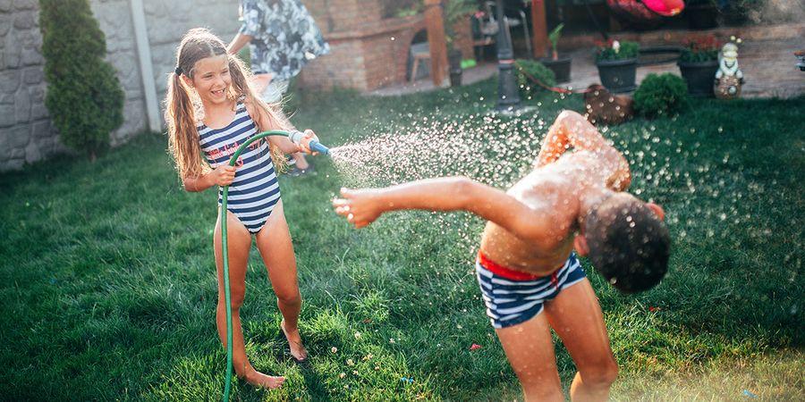 Une fille et un garçon jouent dans le jardin avec un tuyau d'arrosage, un moment amusant de vacances à la maison tiré du guide Photobox pour organiser des vacances à la maison réussies.