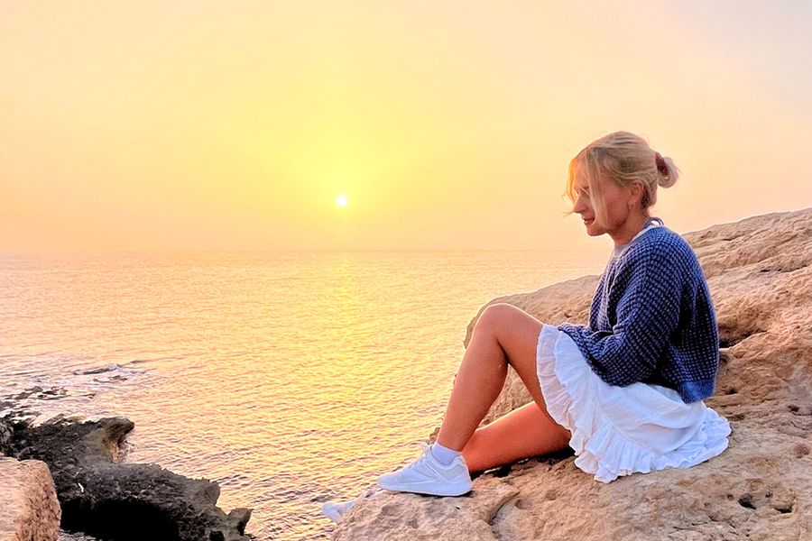 Une femme est assise sur des rochers, regardant la mer et le coucher de soleil, illustrant l'esprit des idées de vacances à la maison aventureuses de Photobox.