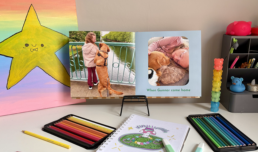 Un album photo enfant ouvert repose sur le bureau d'un enfant entouré de crayons de couleur et de dessins, avec une photo d'une fillette et son chien sur la page de gauche et une autre image accompagnée d'une légende sur la page de droite, illustrant les conseils de Photobox pour associer photos et œuvres d'art dans un album photo familial.