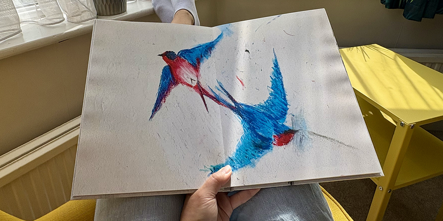 Un album photo enfant ouvert avec une double page pleine d'un dessin d'oiseaux repose sur les genoux d'une femme assise dans un fauteuil, illustrant les étapes de Photobox pour créer un album photo familial à partir des dessins de son enfant.