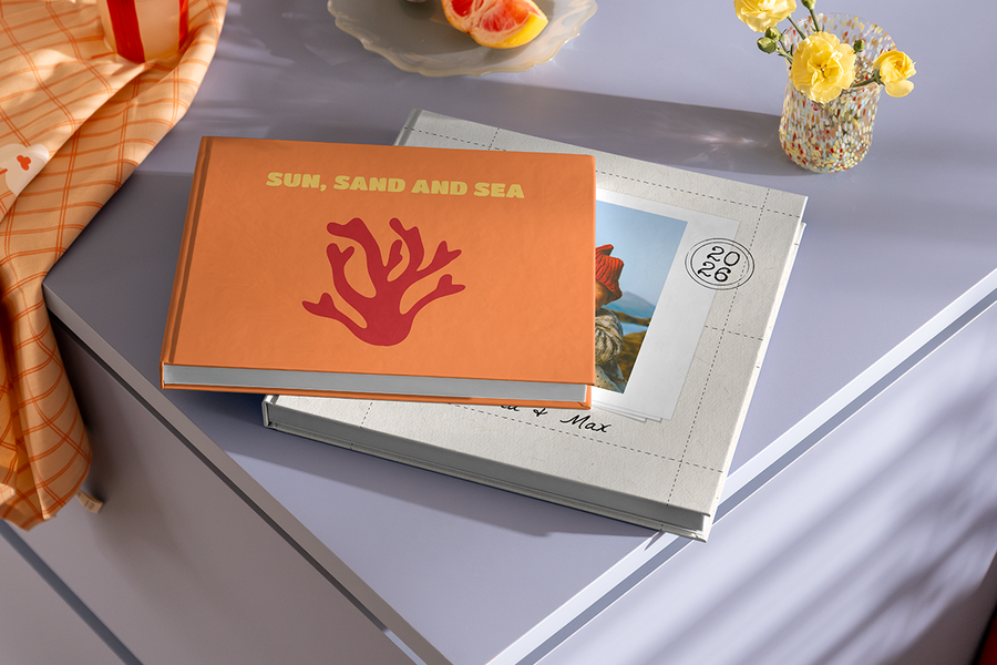 Deux livres photo sont empilés l'un sur l'autre, celui du dessus arborant un motif de corail et le titre 'Sun, Sand and Sea', illustrant les idées de titre album photo vacances à la plage de Photobox.
