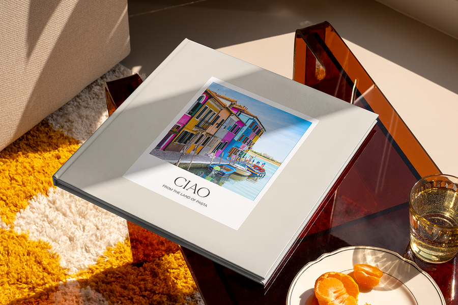 Un livre photo avec un canal et des maisons colorées en Italie sur la couverture s'intitule 'Ciao from the Land of Pasta', illustrant les idées de titre album photo voyage en ville de Photobox.