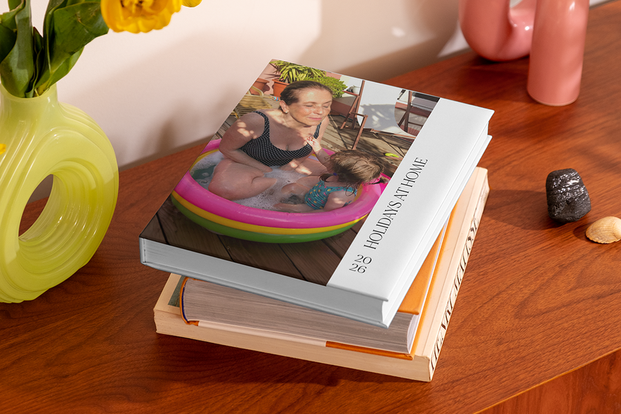Une pile de livres photo comprend un livre du dessus montrant une femme et un jeune enfant dans une piscine gonflable, intitulé 'Holidays at Home', illustrant les idées de titre album photo vacances à la maison de Photobox.