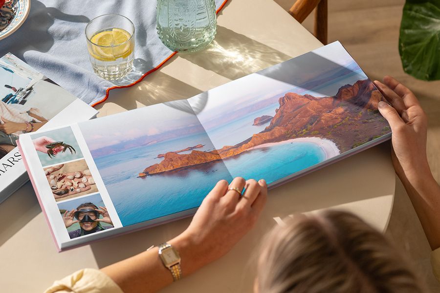 Un livre photo de voyage ouvert sur une table présente une double page avec un magnifique paysage de plage accompagné de trois photos d'été, illustrant le design The Travel Chapter de la nouvelle Summer Travel Series de Photobox.