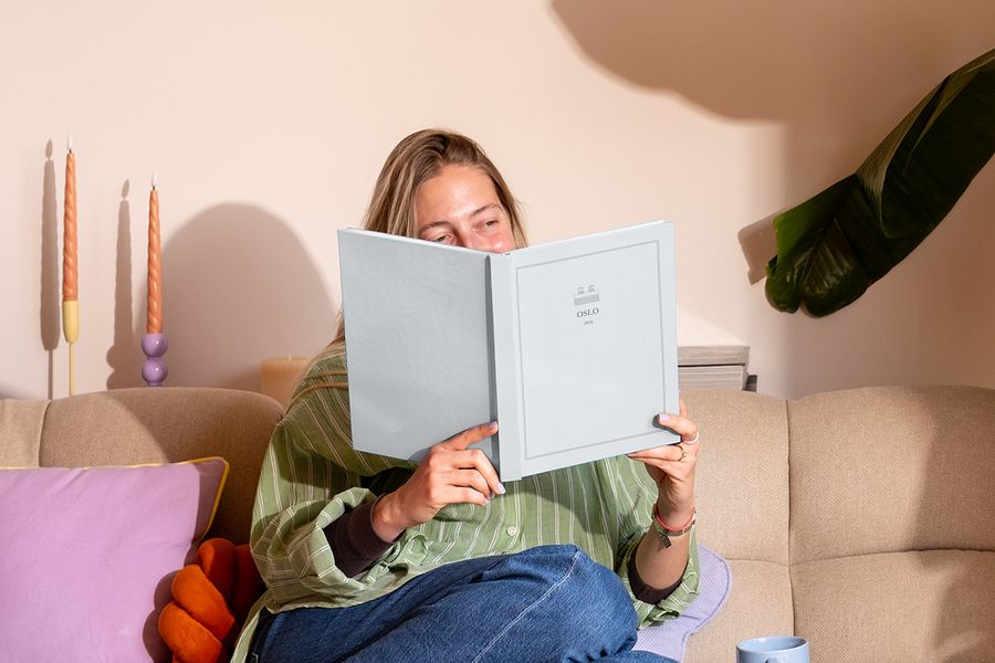 Une femme est assise sur le canapé près d'un coussin coloré, en train de regarder un livre photo de voyage personnalisé intitulé 'Oslo', présentant les nouvelles créations de la Summer Travel Series de Photobox.