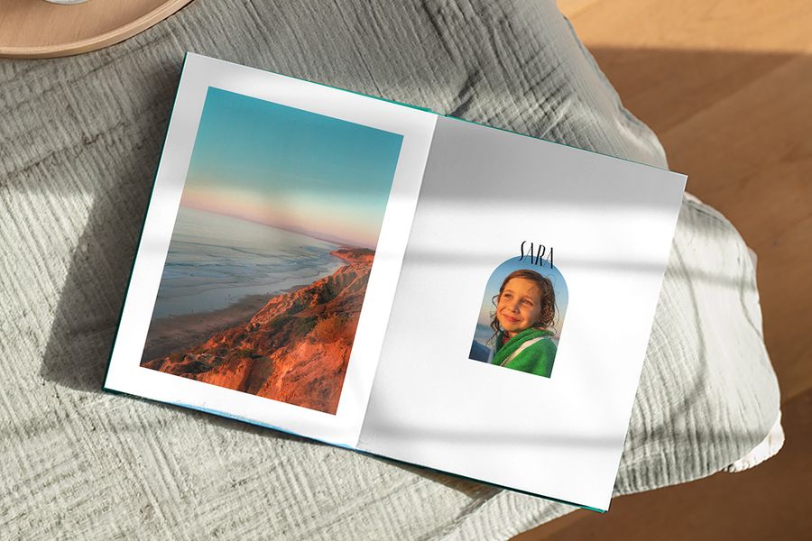 Un livre photo ouvert présente un magnifique paysage sur une page et une photo d'une jeune fille avec la légende 'Sara' sur l'autre, illustrant le design Artisan Masking de Photobox avec des cadres ludiques inspirés des cartes postales et timbres vintage.