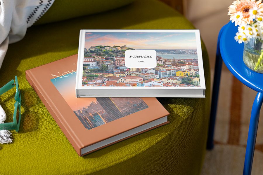 Deux livres photo personnalisés sont empilés sur un tabouret, celui du dessus intitulé 'Portugal' avec une photo du Portugal, et celui du dessous intitulé 'New York' avec une photo de la ville, illustrant les designs personnalisables de la Summer Travel Series de Photobox.