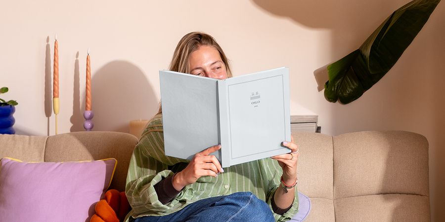 Une femme est assise sur le canapé près d'un coussin coloré, en train de regarder un livre photo de voyage personnalisé intitulé 'Oslo', présentant les nouvelles créations de la Summer Travel Series de Photobox.