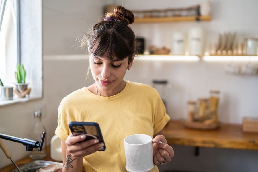 Une femme dans sa cuisine tient une tasse tout en consultant son téléphone, illustrant les conseils de Photobox pour adopter des habitudes de tri photo qui durent.