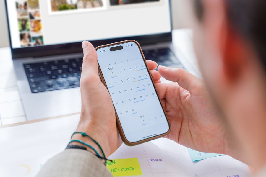Une personne consulte le calendrier sur son téléphone tandis qu'elle travaille sur un livre photo sur son ordinateur portable en arrière-plan, illustrant la routine mensuelle simple de Photobox pour l'entretien des photos.