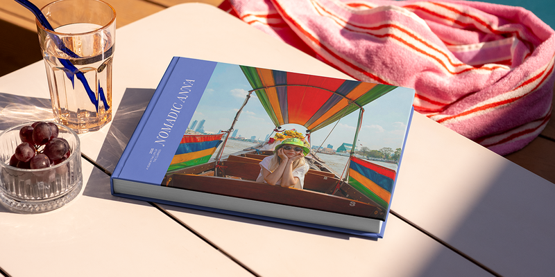 Un grand livre photo de voyage au format paysage, intitulé 'Nomadic Anna', est posé sur une table, avec la photo d'une jeune fille en voyage sur la couverture, illustrant les idées de titre album photo voyage de Photobox.