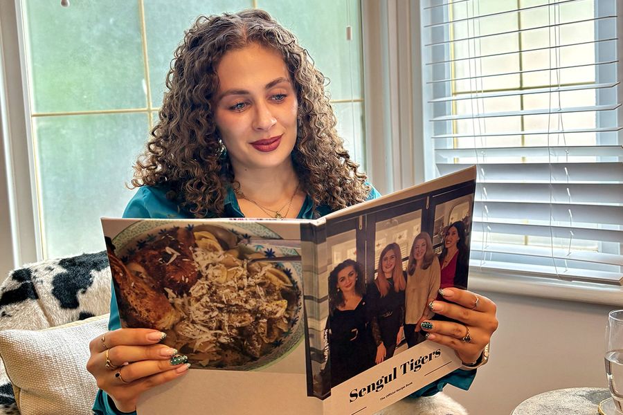 Sarah, qui travaille chez Photobox, est assise dans un fauteuil imprimé peau de vache et regarde son livre photo recette personnalisé, dont la couverture montre quatre femmes, dont Sarah elle-même, illustrant comment un livre de recettes personnalisé réunit recettes et souvenirs de famille.
