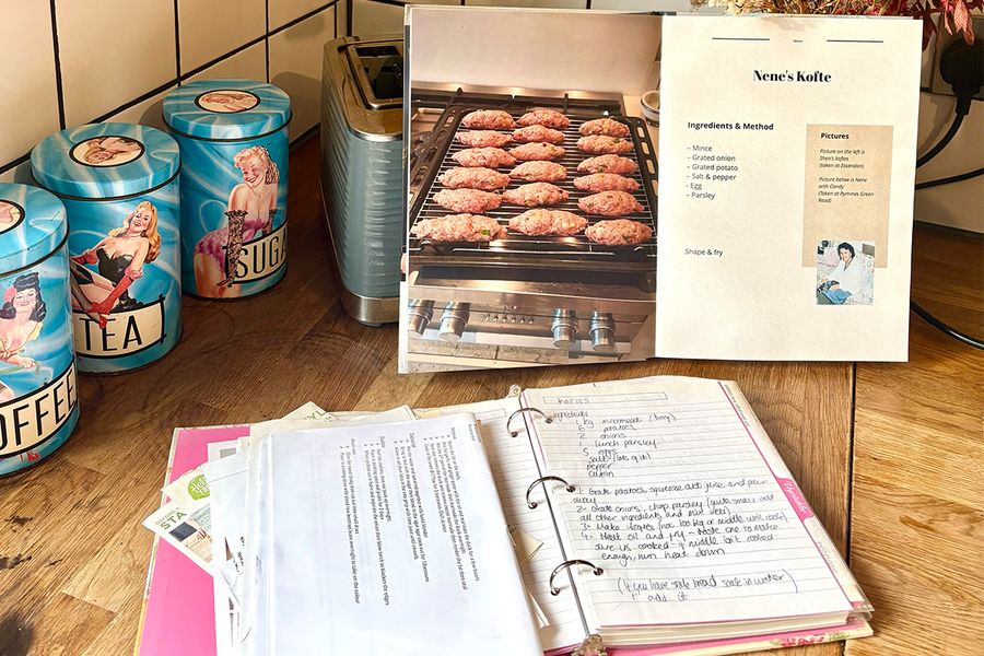 Le livre photo recette personnalisé de Sarah, ouvert sur une recette de kofte de sa Nene, est posé devant un vieux classeur ouvert révélant des recettes familiales manuscrites, les deux reposant sur un plan de travail de cuisine, illustrant comment elle a réuni plusieurs générations de recettes dans un seul livre.