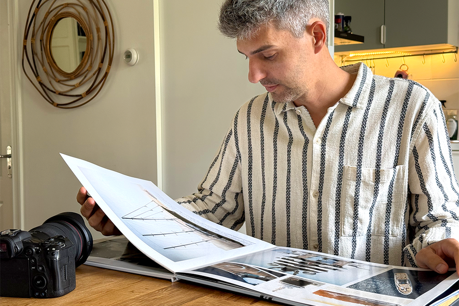 Charl van Rooy est assis dans sa cuisine, regardant son portfolio ouvert, avec son appareil photo posé à côté de lui sur la table, illustrant le guide de Photobox pour créer un portfolio photo sous forme de livre.