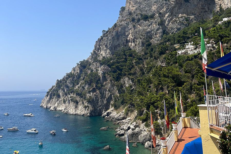 Un littoral méditerranéen avec des collines surplombant une mer bleue parsemée de bateaux, vu depuis un balcon au premier plan, illustrant le charme d'des vacances en septembre en Europe.