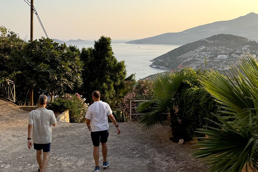 Deux hommes descendent une colline entourée de plantes vers la mer à Kalkan, illustrant le charme des vacances en septembre en Turquie sur la Côte Turquoise.